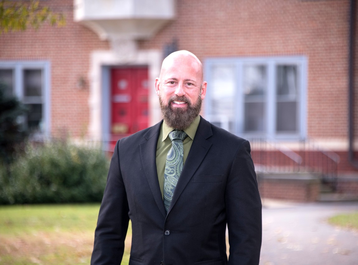 A bearded man in a blazer stands in front of a brick building.
